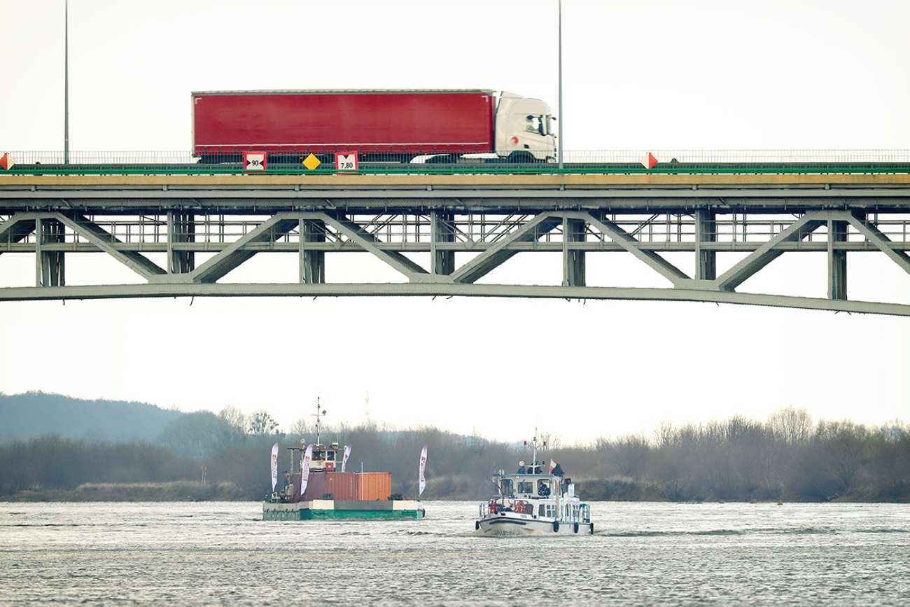 Widok na samochód ciężarowy jadący mostem nad Wisłą. Poniżej płynie barka i mały statek.