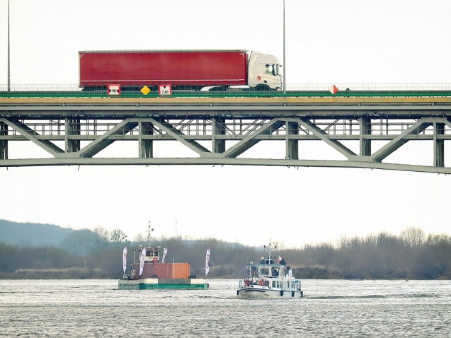 Widok na samochód ciężarowy jadący mostem nad Wisłą. Poniżej płynie barka i mały statek.
